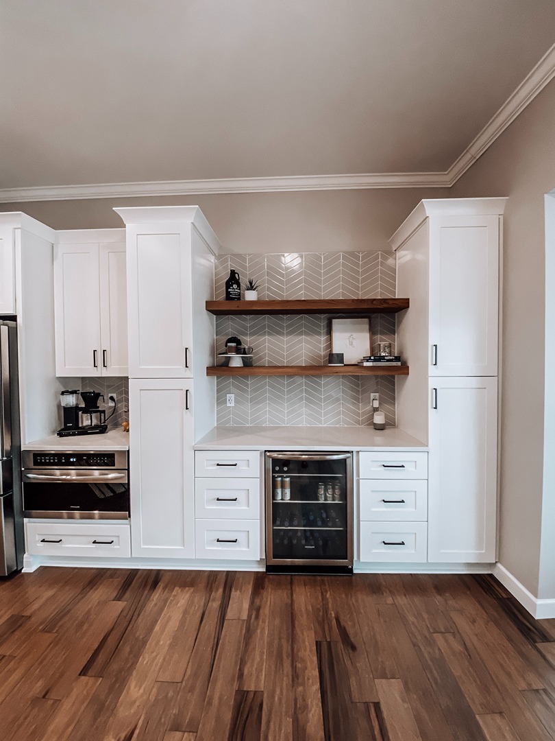 A white coffee bar and beverage centre with floating walnut shelves, a chevron tile backsplash and an integrated drinks fridge